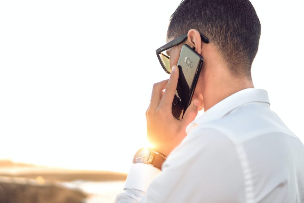 Man in a white shirt holding a smartphone to his ear outdoors, back view with bright sunlight in the background