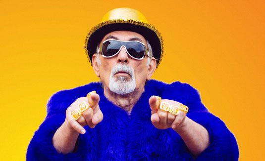 Older man wearing sunglasses and a yellow hat, pointing at the camera against a bright yellow background