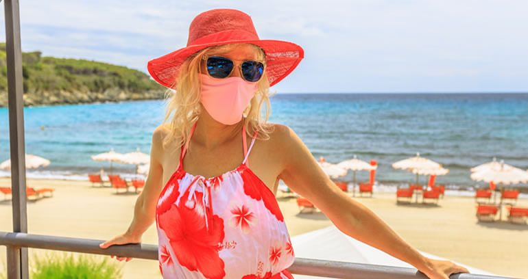 Woman in a red sunhat wearing sunglasses and a pink face mask, standing on a balcony overlooking a beach with sun loungers and parasols