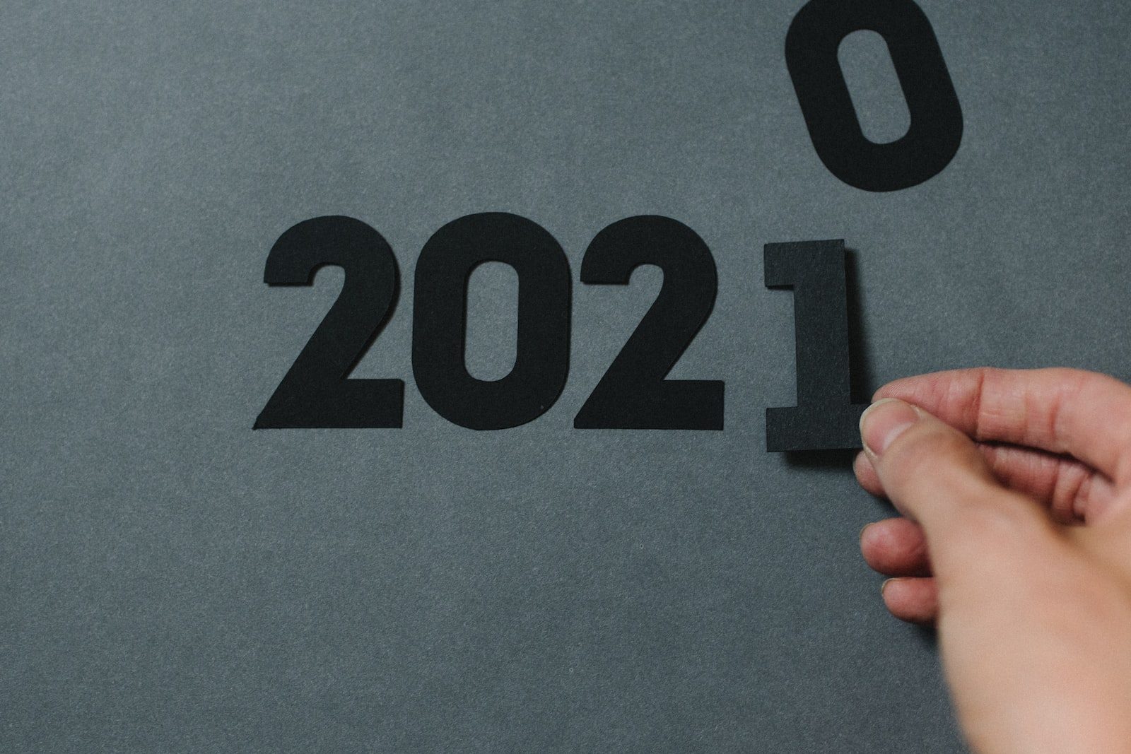 Hand placing a black number 1 next to “2020” to form “2021” on a grey background