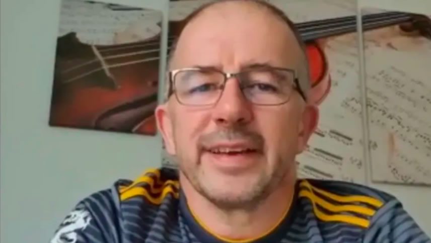 Close-up of a man wearing glasses speaking to camera indoors, with framed wall art behind him