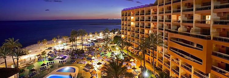 Seafront resort hotel at dusk with illuminated balconies, swimming pool, palm trees and sun loungers beside the shoreline and sea