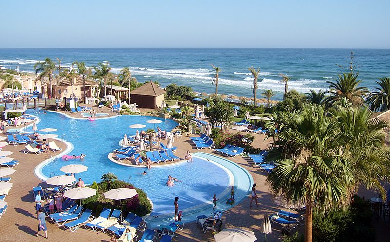 Oceanfront resort swimming pool with sun loungers, parasols and palm trees overlooking the sea in Spain