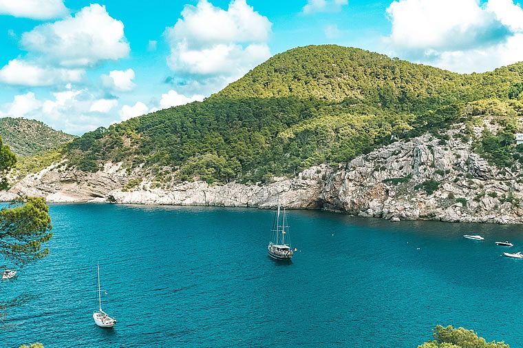 Sailboats anchored in a turquoise bay below rocky cliffs and green hills under a blue sky with scattered clouds