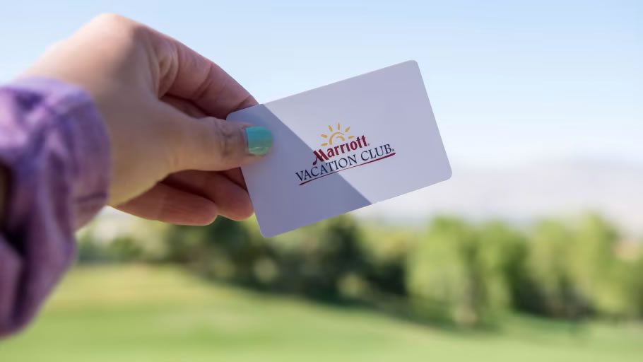 Hand holding a Marriott Vacation Club card against a blurred outdoor resort backdrop and blue sky