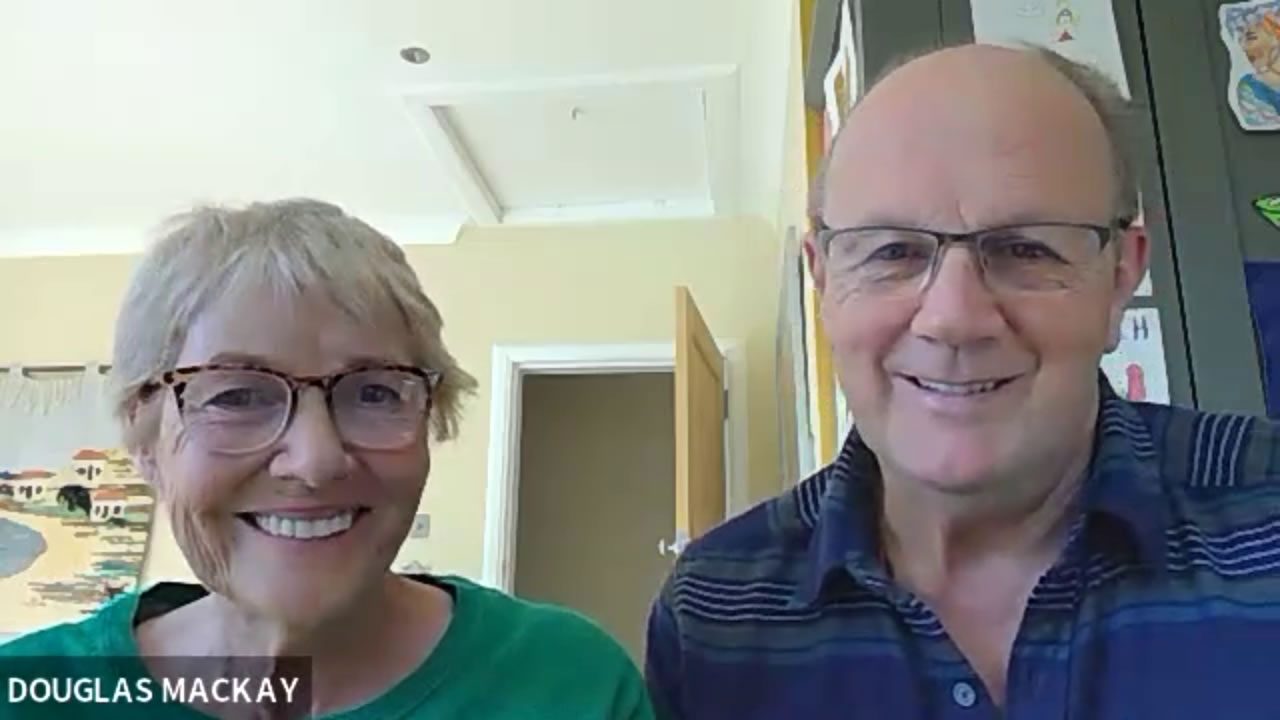 Douglas and Linda Mackay smiling at the camera during a video call, seated indoors at home with an open door behind them