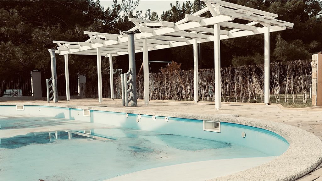 Empty outdoor swimming pool beside a pergola walkway and closed gate at a quiet resort, suggesting timeshare resort facilities being reduced