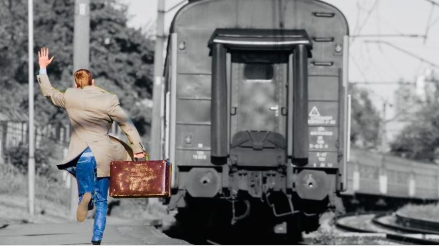 Man with suitcase running alongside a departing train on a railway platform, waving as it pulls away