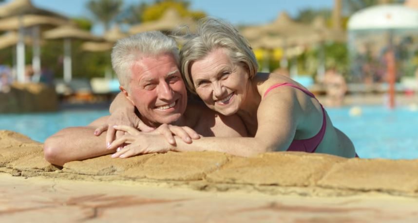 Smiling older couple relaxing at the edge of a resort swimming pool with sun loungers in the background