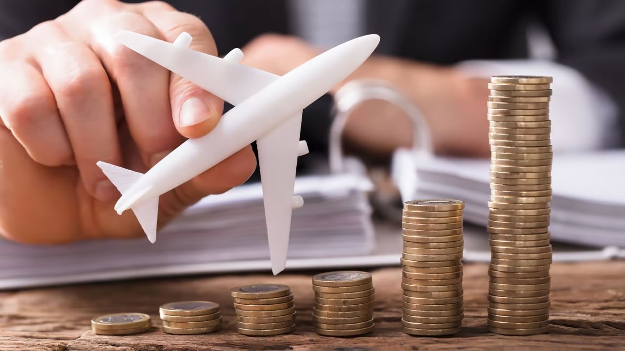Hand holding a model aeroplane next to stacks of coins increasing in height, showing rising travel costs and fees