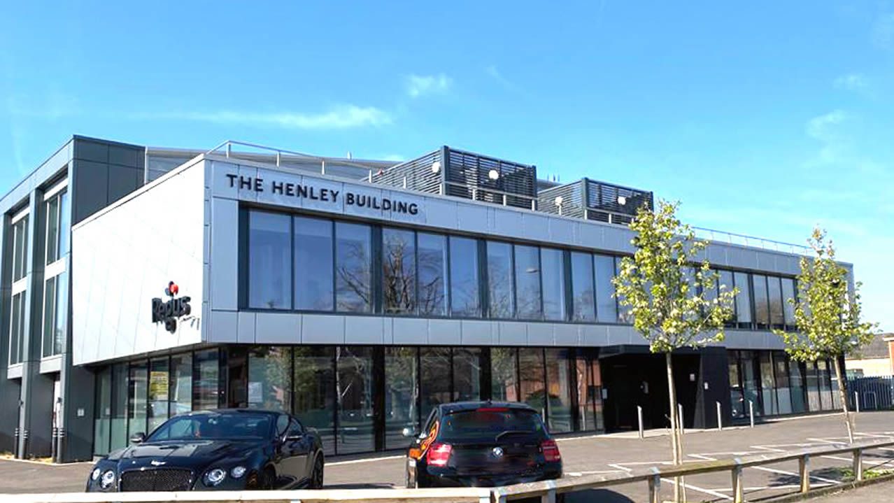 Exterior of The Henley Building office block with large glass windows, parked cars and young trees in front under a blue sky
