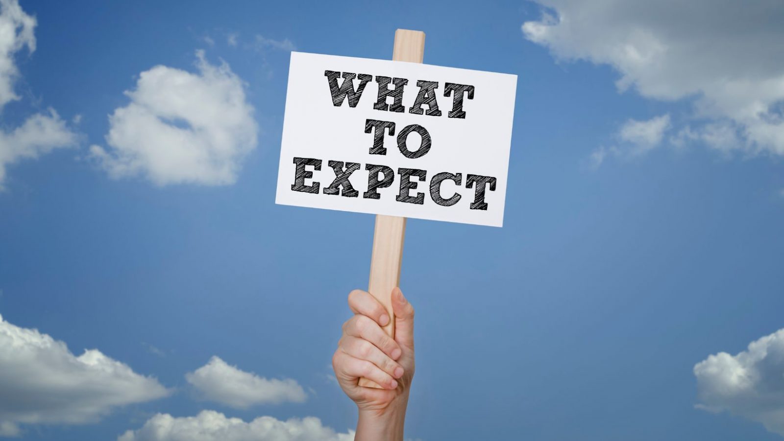 Hand holding a wooden sign reading “What to expect” against a blue sky with clouds