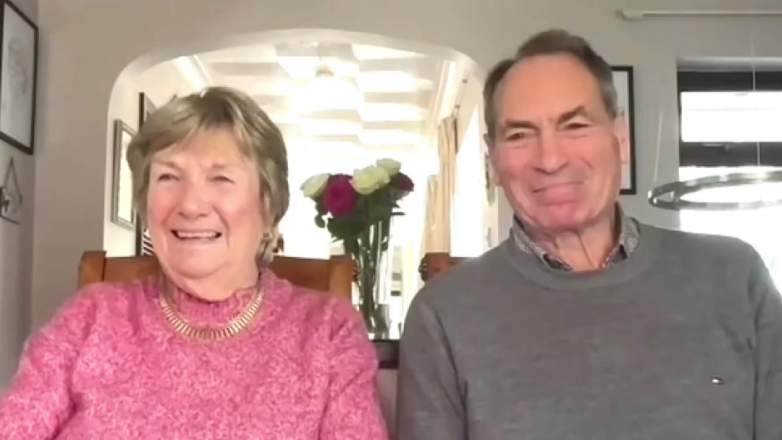 Smiling older couple sitting indoors in a bright room, with a vase of flowers behind them during a video call interview about a timeshare claims win story