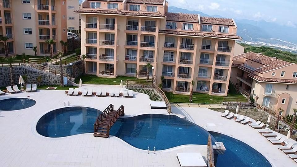 Large apartment-style resort complex with an outdoor swimming pool, sun loungers and parasols on a terrace with hills in the background