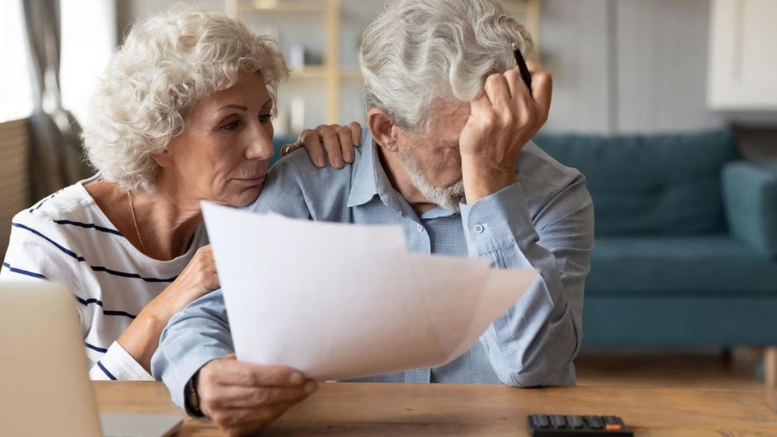 Worried elderly couple at a table reading paperwork, with the man holding his head in his hand as the woman comforts him in the background