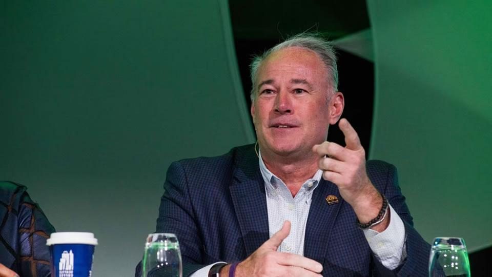 Man in a suit speaking and gesturing at a table with water bottles and a takeaway coffee cup against a green background