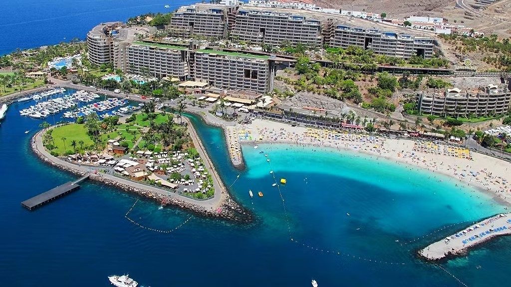 Aerial view of a large coastal resort with multi-storey hotel buildings, marina, palm-lined grounds and a crowded sandy beach beside a turquoise bay