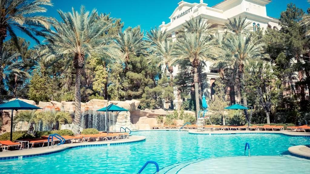 Outdoor hotel pool with sun loungers and palm trees, with a resort building in the background displaying “JW Marriott” signage