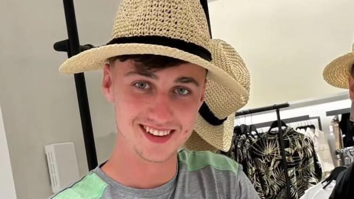 Young man smiling in a straw hat inside a clothing shop, with hanging hats and patterned garments behind him