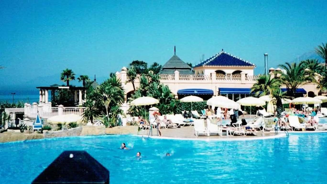 Sunlit resort swimming pool with people relaxing on loungers and parasols beside a Mediterranean-style hotel building and palm trees by the sea