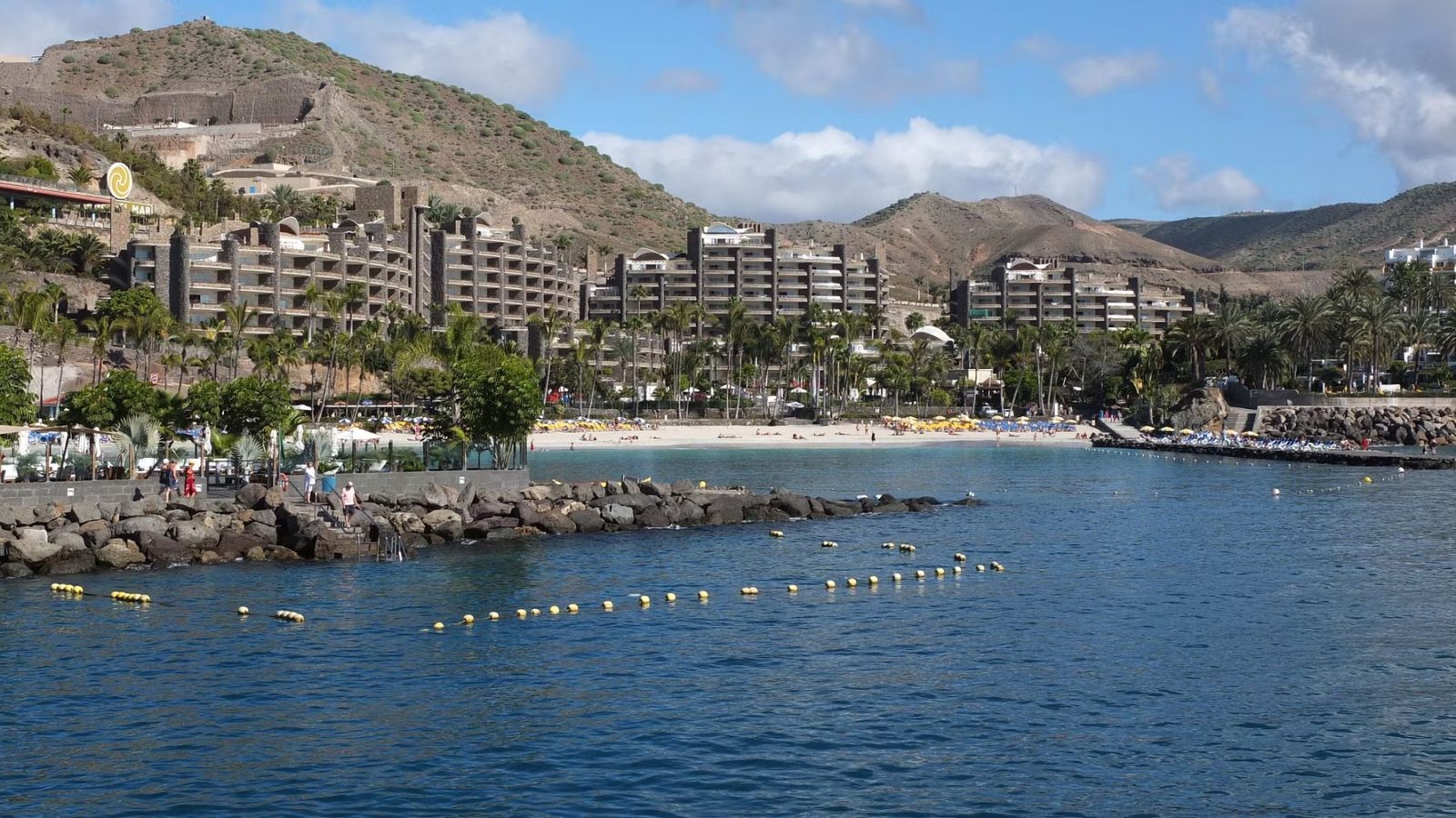 Coastal holiday resort apartments on a hillside behind a sandy beach and calm bay with palm trees and sunloungers