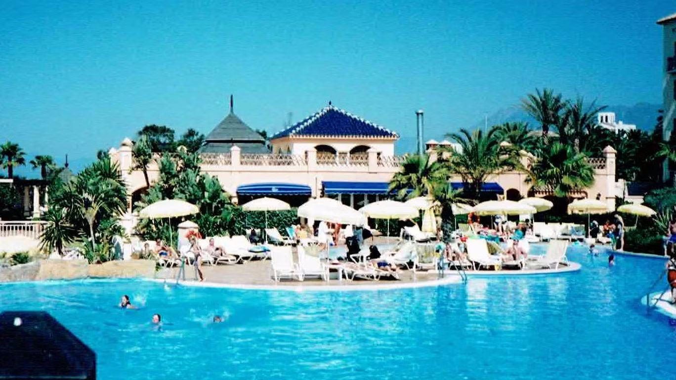 Outdoor hotel pool with sun loungers and parasols in front of a Spanish-style resort building with palm trees and mountains in the background