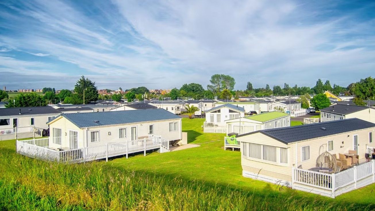 Static caravan holiday park with rows of white lodges on green lawns under a cloudy blue sky