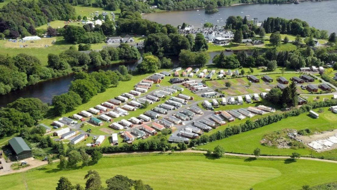 Aerial view of a UK holiday park with rows of static caravans beside a river and lake, surrounded by trees and green fields