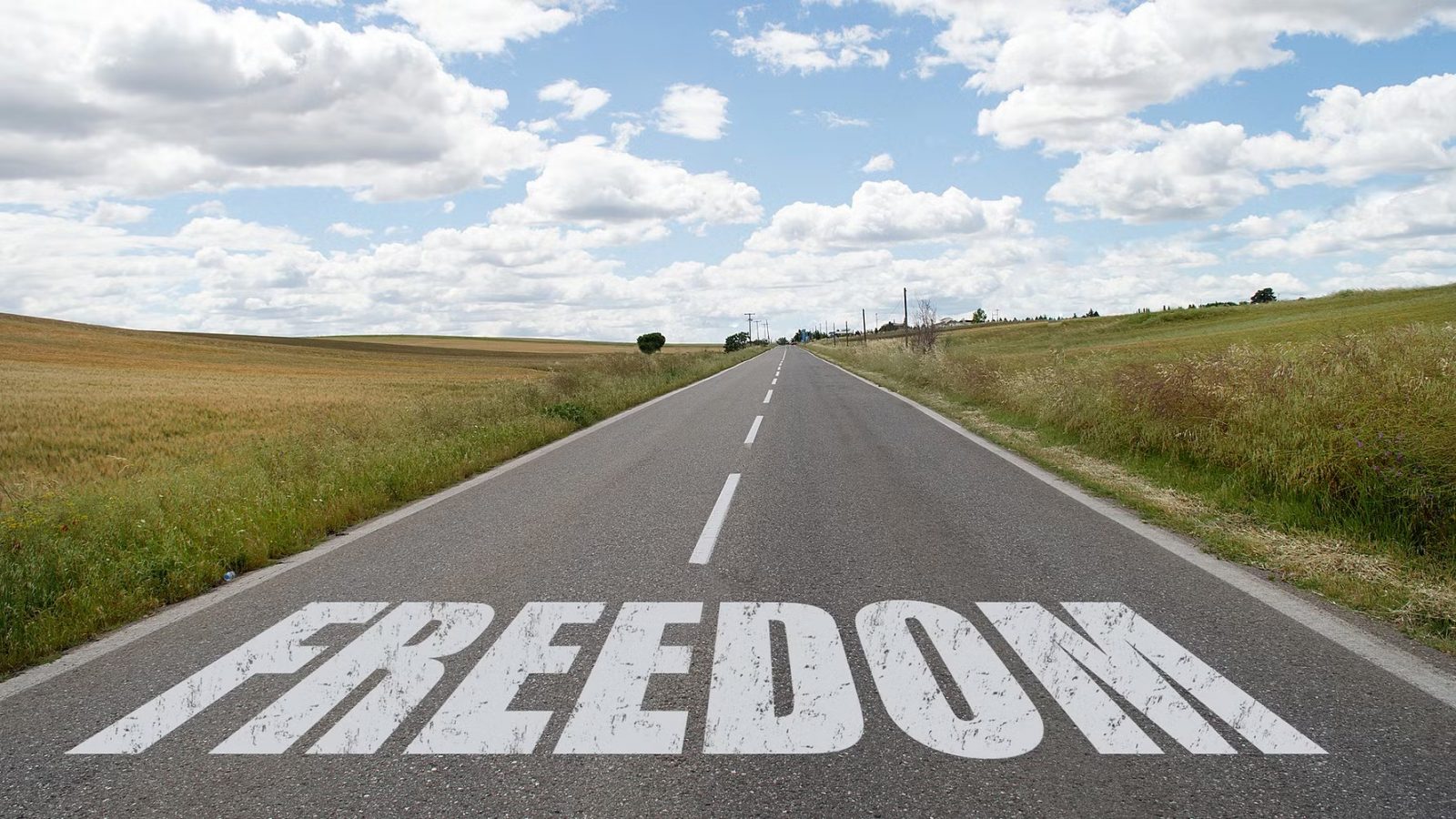 Open road through countryside with the word “FREEDOM” painted in large white letters on the tarmac under a cloudy sky