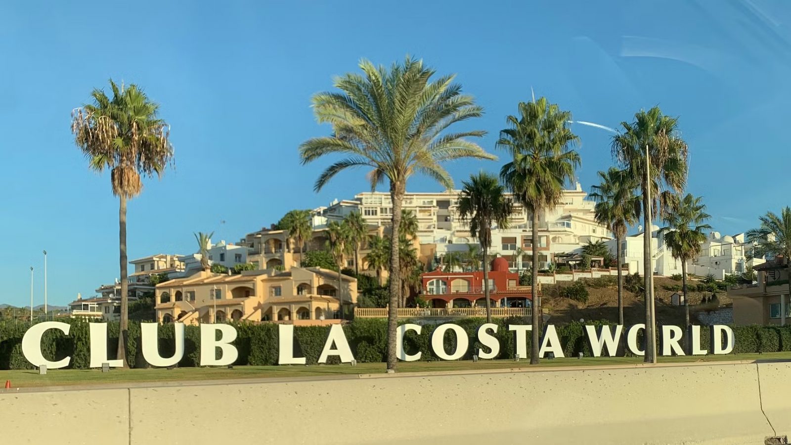 Entrance sign reading “CLUB LA COSTA WORLD” in front of a palm-lined resort complex under a clear blue sky