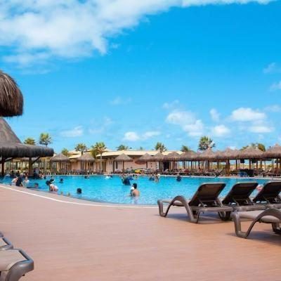 Sun loungers beside a large outdoor resort swimming pool with people in the water, palm trees and a blue sky in the background