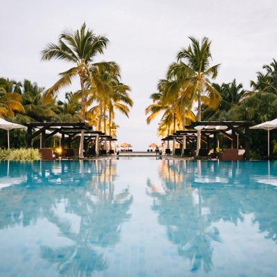 Infinity swimming pool lined with palm trees, sun loungers and parasols leading towards the sea at sunset