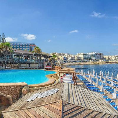 Seaside resort in Malta with a curved swimming pool, wooden sun deck and rows of blue loungers and white parasols beside the sea under a clear sky