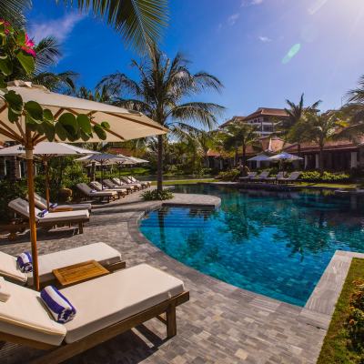Sun loungers and parasols beside a tropical resort swimming pool with palm trees and hotel buildings in the background