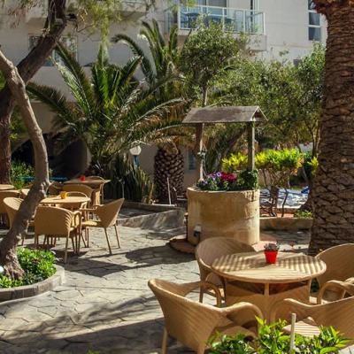 Outdoor courtyard seating area with wicker tables and chairs, palm trees, a stone well planter and a swimming pool beside an apartment building