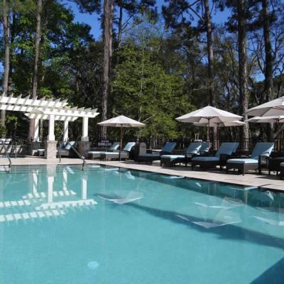Outdoor resort swimming pool with sun loungers and white parasols beside a pergola, surrounded by tall trees