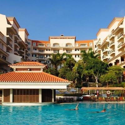 Outdoor swimming pool with loungers in front of the Madeira Regency Palace hotel building under a clear blue sky