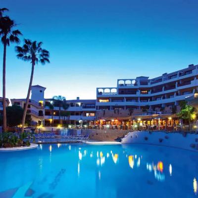 Evening view of a resort hotel with palm trees and a lit swimming pool in the foreground under a clear blue sky