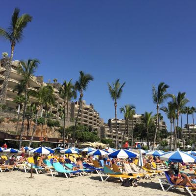 Crowded beach with sun loungers and blue-and-white parasols in front of a large seaside resort complex and tall palm trees under a clear blue sky