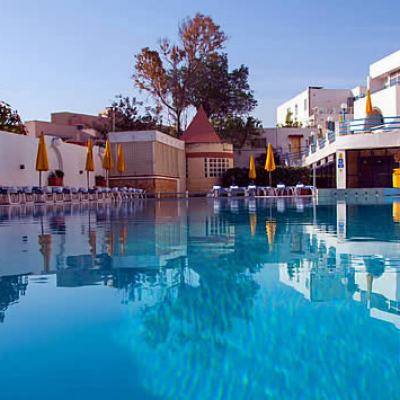 Outdoor swimming pool with sun loungers and closed parasols beside white resort buildings under a clear blue sky