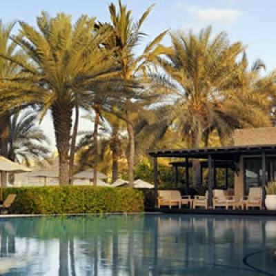 Resort swimming pool with sun loungers and palm trees beside a low modern building in warm evening light