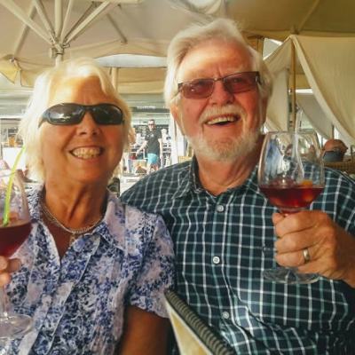 Smiling older couple wearing sunglasses raise glasses of red wine while seated under parasols at an outdoor café terrace