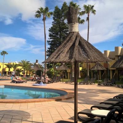 Outdoor swimming pool with sun loungers and thatched parasols in a yellow resort complex with palm trees under a blue sky in Fuerteventura