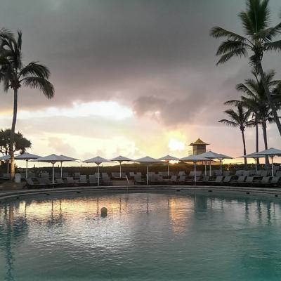Sunset over a resort swimming pool with palm trees, sun loungers and parasols under dark clouds