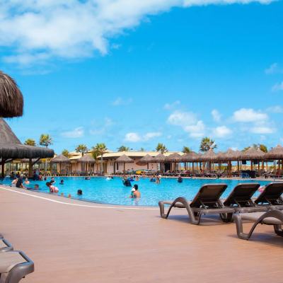 Sunlit resort swimming pool with people in the water, rows of sun loungers and thatched parasols under a blue sky