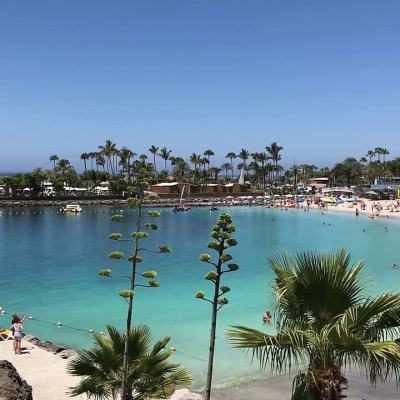 Crowded beach and turquoise lagoon with sun loungers, palm trees and a large resort building in the background under a clear blue sky