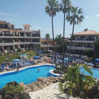 Outdoor hotel pool surrounded by sun loungers, parasols and palm trees between multi-storey apartment buildings on a sunny day
