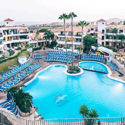 Aerial view of the Parque Albatros resort with a large swimming pool, rows of blue sun loungers, palm trees and white apartment buildings with terracotta roofs.
