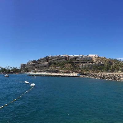 Coastal resort skyline overlooking a calm blue sea, with a rocky breakwater, palm trees and boats under a clear sky