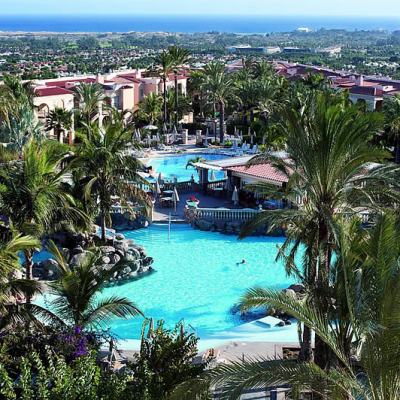 Palm-lined resort swimming pool with sun loungers and terracotta-roofed buildings, overlooking Maspalomas and the sea in Gran Canaria